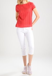Červený top s krátkým rukávem a květinovým výřezovým vzorem, spárovaný s bílými zkrácenými kalhotami a světle růžovými teniskami. Hladký povrch, ležérní střih.