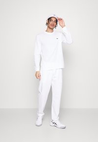 Chemise blanche à manches longues et pantalon assorti en coton. Détail logo vert discret sur la poitrine. Une casquette blanche et des chaussures complètent la tenue.