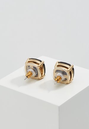 Boucles d'oreilles clous en or avec des pierres carrées noires et des attaches en plastique transparent reposant sur un cube blanc sur un fond gris clair.