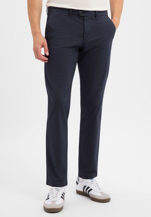 Man wearing slim-fit dark blue trousers and white sneakers with black stripes standing with one hand in pocket.