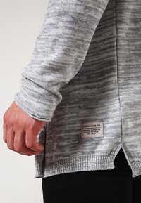 Ljust grå, stickad tröja med ett strukturerat mönster, ribbad nederkant och sidoklyftor. Har en tygetikett med varumärket på nedersta vänstra sidan.
