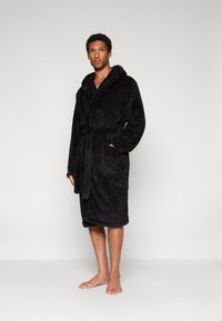 Black, plush bathrobe featuring a hood, tie belt, and two side pockets; soft texture with hemmed edges. Model standing barefoot.
