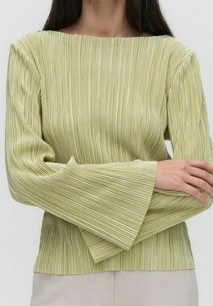 Mujer con una blusa plisada de manga larga color verde claro con puños anchos, combinada con pantalones de color claro, con los brazos cruzados a la altura de la cintura.