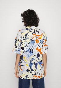 Osoba z kręconymi włosami stoi tyłem, ubrana w koszulę z krótkim rękawem w abstrakcyjny kwiatowy wzór w kolorach niebieskim, pomarańczowym, żółtym i czarnym.