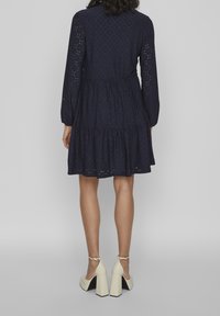 Mörkblå klänning med långa ärmar, texturerad ögla tyg, flerfaldig volangnederdel och nyckelhålsdetalj i ryggen. Matchad med crèmefärgade klackskor.