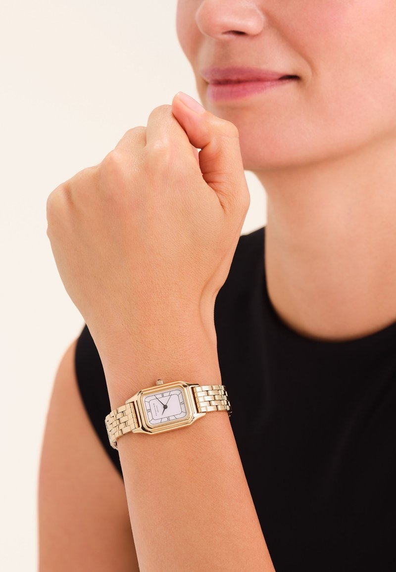 Reloj de pulsera de metal dorado con una esfera blanca rectangular, manecillas negras y una pulsera texturizada. Lleva puesto en una muñeca, contra un fondo simple.