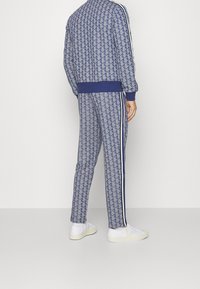 En person bär en blå mönstrad träningsdräkt med vita sidostråkar och vita sneakers, stående mot en neutral ljus bakgrund, vänd bort.