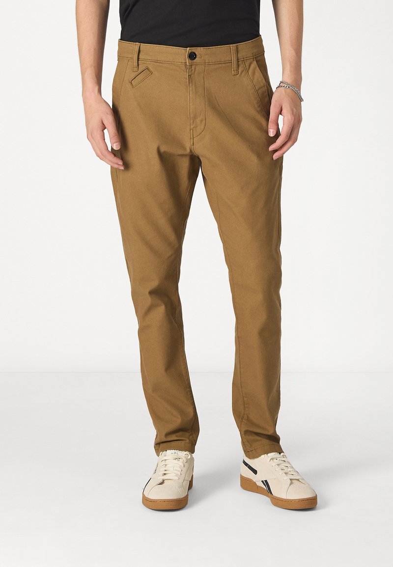 Tan bomullbyxor med rak benstil, med framfickor, bältesöglor och en enkel knäppning, bärs tillsammans med vita sneakers.