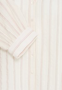 Poignet de chemise blanche et devant boutonné avec des rayures fines verticales rose et beige sur un tissu texturé.