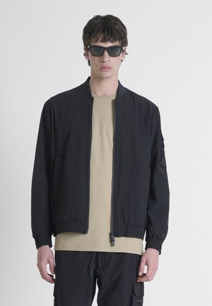 Black bomber jacket with a front zipper, ribbed cuffs, and side pockets, worn over a beige t-shirt; model wearing angular black sunglasses.