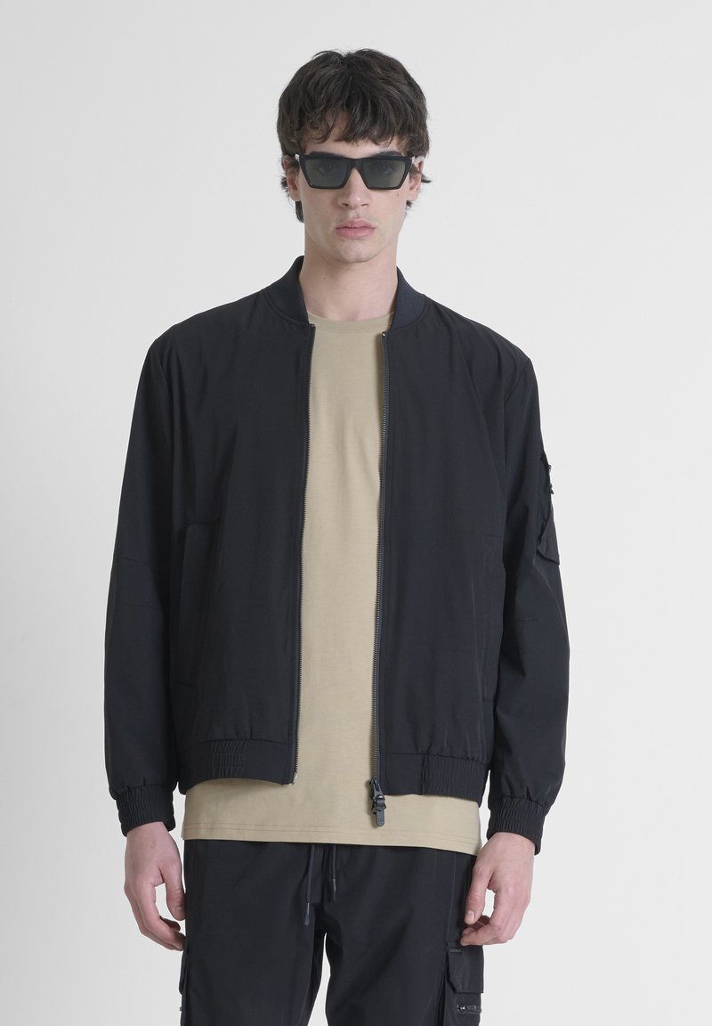 Chaqueta bomber negra con cremallera frontal, puños acanalados y bolsillos laterales, llevada sobre una camiseta beige; modelo con gafas de sol negras angulares.
