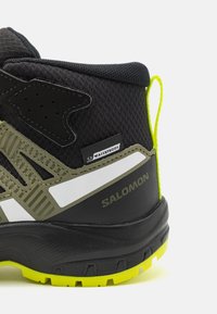 Czarny i oliwkowy but trekingowy z etykietą wodoodporności, teksturowaną cholewką, gumową podeszwą oraz jaskrawożółtymi akcentami na pięcie i podeszwie.