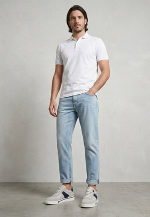 Mand stående med den ene hånd i lommen iført hvid poloshirt, lyseblå op rullede jeans og hvide sneakers med navy detaljer på gråt gulv.