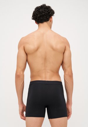 Homme aux cheveux bouclés et foncés vu de dos, portant un boxer noir ajusté, debout devant un fond clair uni.