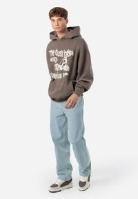Felpa marrone con testo e grafica bianchi, abbinata a jeans azzurri chiari e sneakers marroni e bianche. Il tessuto appare morbido e ha un design comodo.