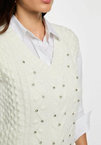 Gilet bianco lavorato a maglia con scollatura a V, caratterizzato da motivi a treccia e decorato con perline argentate. Indossato sopra una camicia bianca con colletto.