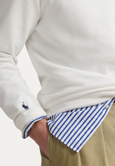 Balta ilgomis rankovėmis marškiniai su mėlynomis dryžuotomis pamušalo detalėmis ir tamsiai mėlynu logotipu ant rankov�ės. Turi klasikinį apykaklę ir švelnų tekstūrą.