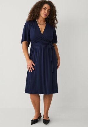 Kobieta o kręconych włosach, ubrana w granatową sukienkę kopertową i czarne baleriny, stojąca na tle jednolitego jasnoszarego tła.