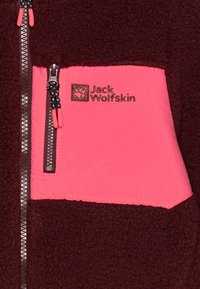 Chaqueta de forro polar marrón con bolsillo con cremallera rosa; cuenta con cremallera negra y logotipo de marca. Textura suave, elementos de color en contraste.