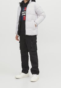 Jasnoszara kurtka puchowa na zamek, czarna bluza z kapturem z logo, czarne spodnie cargo oraz białe sneakersy. Nacisk na funkcjonalność i wygodę.