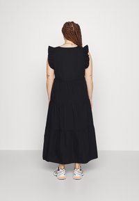 Femme aux cheveux tressés portant une robe midi noire à volants avec des manches à volants et des baskets blanches, debout devant un fond uni.