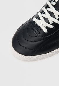 Κοντινή λήψη από το μπροστινό μέρος μαύρου δερμάτινου αθλητικού παπουτσιού με λευκά κορδόνια και υφασμένο λευκό λαστιχένιο πατούμενο σε γκρι φόντο.