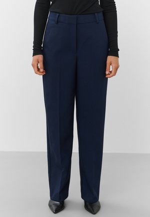 Femme portant un pantalon droit bleu marine ajusté, des chaussures noires pointues et un haut noir à manches longues, debout devant un fond uni.