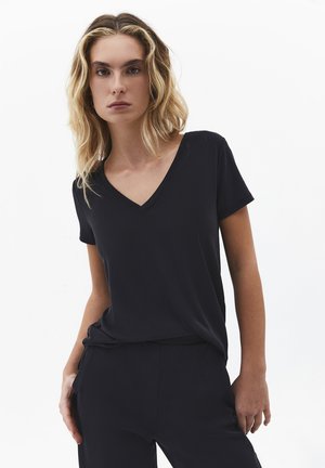 Blonde vrouw met halflang haar, die een zwart V-hals shirt met korte mouwen en een zwarte broek draagt, voor een witte achtergrond.