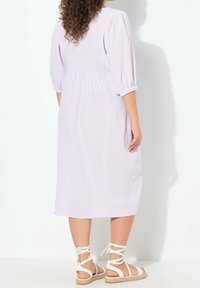 Lila, knielanges Kleid mit puffärmel und geraffter Taille. Getragen mit weißen Riemchensandalen mit Knöchelbindungen und geflochtenen Sohlen.