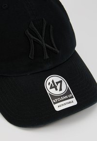 Gorra de algodón negra con el logo bordado "NY" en color a juego. Cuenta con una correa ajustable con una etiqueta circular blanca etiquetada "47 CLEAN UP."