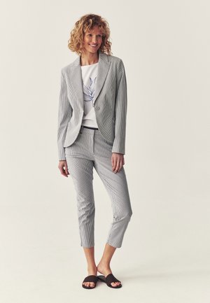 Femme souriante aux cheveux bouclés portant un blazer gris clair à fines rayures, un pantalon court assorti, une chemise blanche à motifs floraux et des sandales slides noires.