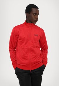 Sarkans zip-up jaka, izgatavota no gludas auduma, ar augstu apkakli. Izceļas ar melnu EA7 logo uz krūtīm un diskrētām sānu kabatām.