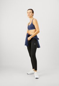 Sujeción deportiva azul marino, leggings negros con franjas blancas a los lados, combinados con una camiseta azul atada a la cintura y zapatillas deportivas blancas.