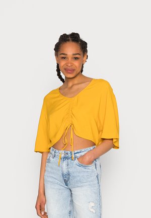 Top cropped amarelo com decote redondo e cordão na frente. Apresenta mangas curtas largas e um tecido leve e texturizado. Combinado com jeans desbotados de cor azul clara.