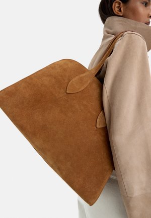 Brązowa torba typu tote z zamszu o dużym trójkątnym kształcie, z dwoma zakrzywionymi uchwytami i gładką fakturą, noszona na beżowej odzieży.