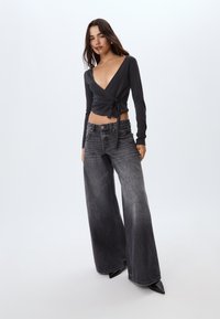 Top nero avvolgente con scollatura a V profonda, legato in vita; abbinato a jeans grigi sbiaditi a gamba larga; modello con tacchi neri a punta.