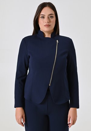Veste bleu marine avec un col montant, dotée d'une fermeture éclair diagonale en or et d'une coupe ajustée. Texture lisse, manches longues et détails minimalistes.