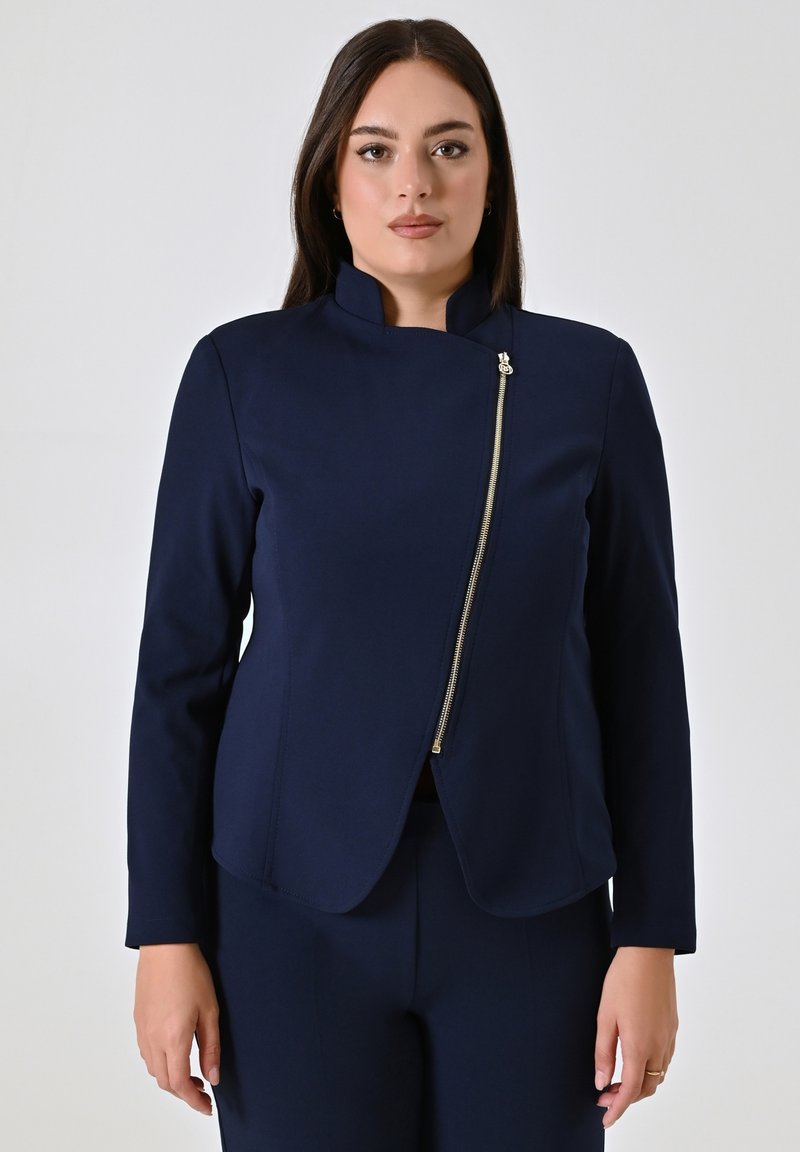 Marineblauwe jas met een hoge kraag, voorzien van een diagonale gouden rits en een getailleerde pasvorm. Glad textuur, lange mouwen en minimale details.