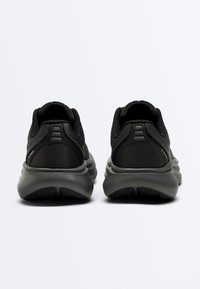 Paire de baskets de sport noires vues de dos, avec des dessus en tissu texturé et des semelles épaisses grises rembourrées, sur fond blanc.