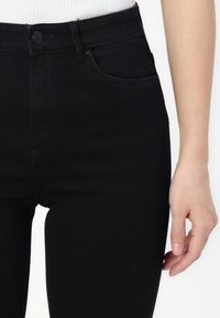 Jeans skinny negros con una cintura alta, cierre frontal de botón, cinco bolsillos y una textura de denim suave y elástica.
