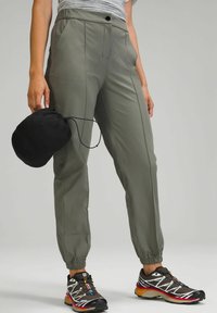 Grijze sportbroek met een elastische tailleband, taps toelopende boorden en zichtbare stikseldetails. In één hand wordt een zwarte tas vastgehouden.