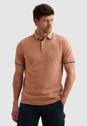 Mężczyzna w jasnobrązowej koszulce polo z czarnym obszyciem i wypukłym napisem na piersi i rękawach, stoi z rękami w kieszeniach.