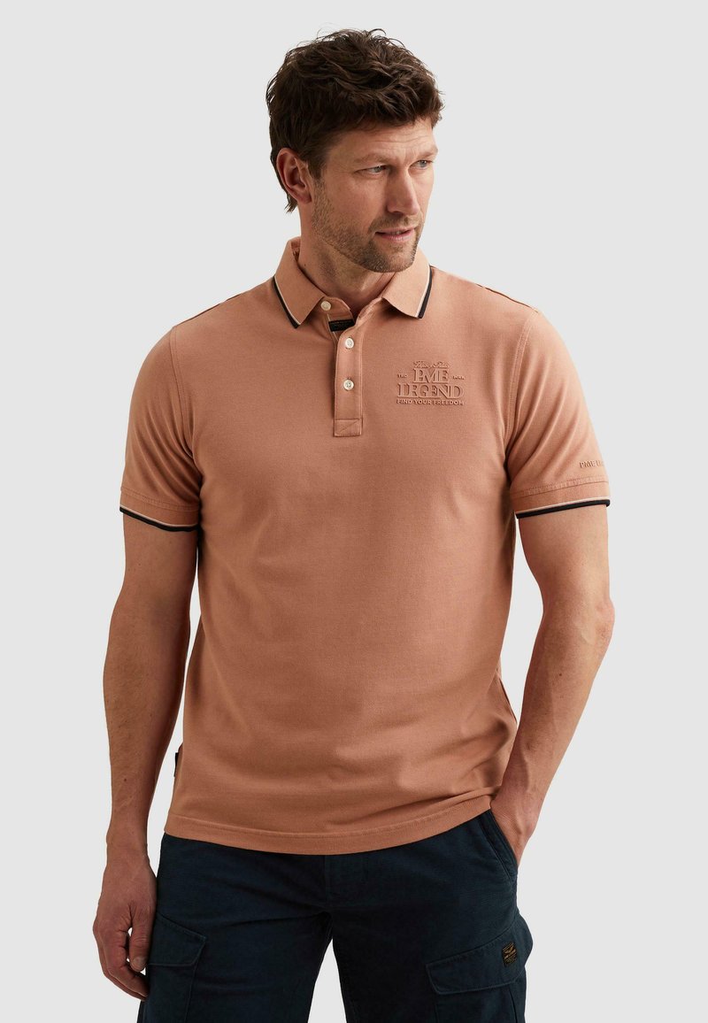 Homme portant un polo marron clair avec un liseré noir et un texte en relief sur la poitrine et les manches, debout les mains dans les poches.