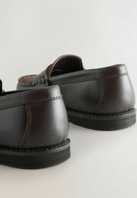 Ein Paar dunkelbraune Lederloafers mit schwarzen Gummisohlen, von hinten auf einer hellen Oberfläche gezeigt.