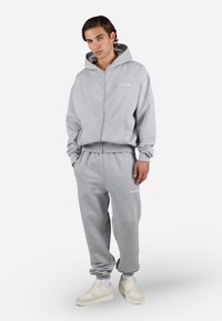 Grijze rits-hoodie met voorzakken, gecombineerd met bijpassende joggingbroek. Zacht materiaal, geribbelde manchetten en witte logo-detail op de broek.