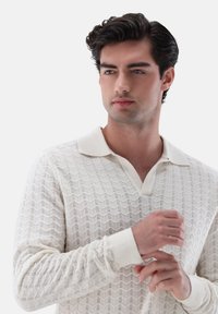 Junger Mann in einem strukturierten weißen Langarmshirt mit Kragen, der sein Manschette anpasst und leicht zur Seite schaut.