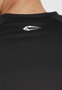 Schwarzes Sportshirt aus strukturiertem Stoff, mit rundem Ausschnitt und einem kleinen weißen Logo am hinteren Halsbereich. Nahtdetails sind sichtbar.