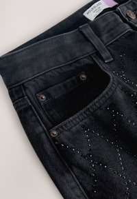 Schwarze Jeans aus Denim mit Fronttasche, Gürtelschlaufen, metallischen Nieten und dezenten sternförmigen schwarzen Perlenverzierungen auf dem Stoff.