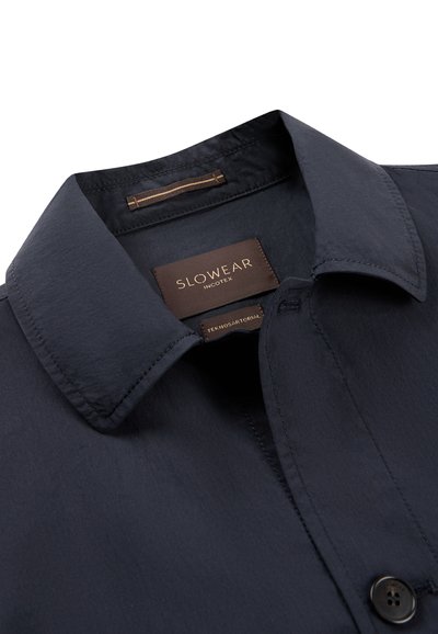 Col de chemise bleu marine foncé avec une étiquette marron visible portant l'inscription "SLOWEAR INCOTEX" et une petite étiquette "TEKNOSARTORIAL" à l'intérieur de l'encolure.