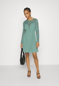 Um vestido azul-petróleo com corpete justo, mangas longas em renda e saia rodada. Apresenta um decote em forma de V e é combinado com sapatos de salto com tiras e uma bolsa preta.